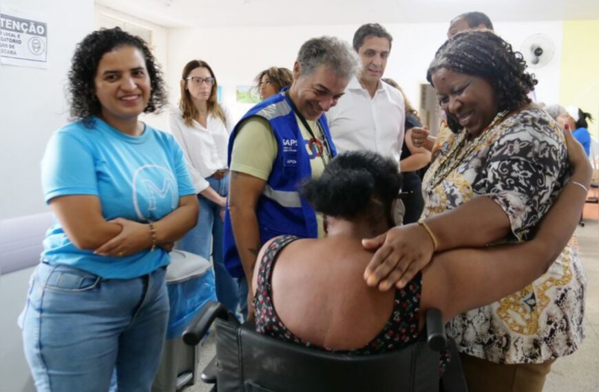 Mulher em cadeira de rodas recebe abraço carinhoso em ambiente de posto de saúde, cercada por pessoas sorridentes.