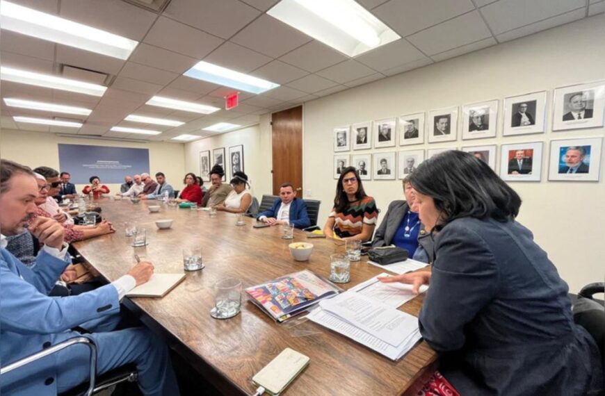 Reunião formal com várias pessoas sentadas ao redor de uma mesa grande, todas atentas.