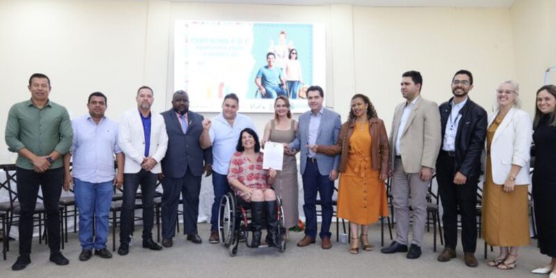 Grupo diverso posando sorridente, com mulher em cadeira de rodas ao centro segurando um documento.