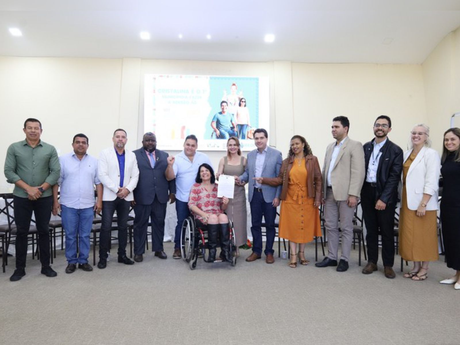 Grupo diverso posando sorridente, com mulher em cadeira de rodas ao centro segurando um documento.
