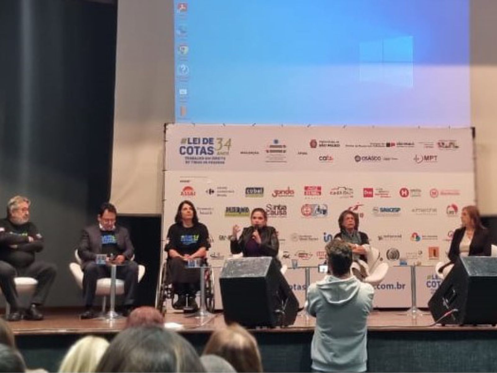 Mesa de debate sobre Lei de Cotas com seis pessoas no palco e plateia assistindo.