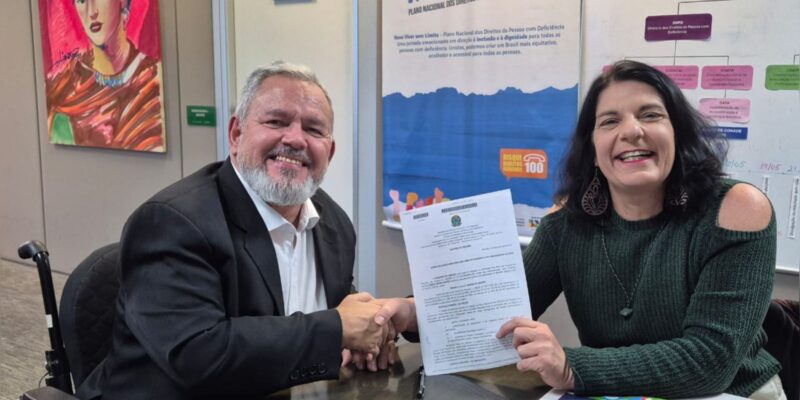 Homem e mulher sorrindo, apertam as mãos e exibem documento oficial em escritório colorido.