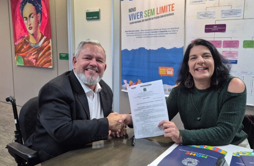 Homem e mulher sorrindo, apertam as mãos e exibem documento oficial em escritório colorido.