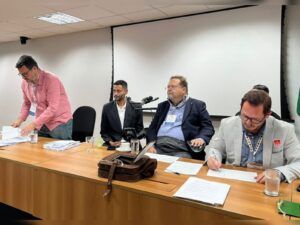 Comissão do Conade delibera por nova eleição para vagas da sociedade civil no mandato 2025 2028 2 Mesa de reunião com cinco homens, alguns sentados e um em pé, todos de roupa social. Há papéis, copos e uma bolsa sobre a mesa. Ao fundo, tela de projeção branca.