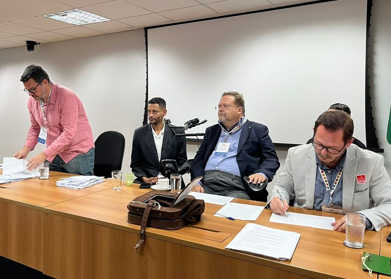 Mesa de reunião com cinco homens, alguns sentados e um em pé, todos de roupa social. Há papéis, copos e uma bolsa sobre a mesa. Ao fundo, tela de projeção branca.