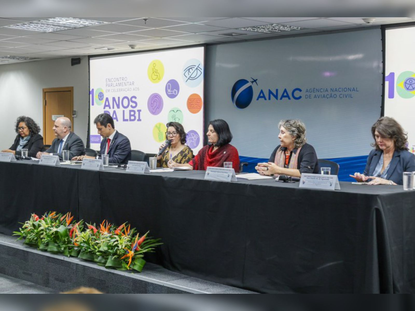 Mesa de conferência da ANAC com sete pessoas e cartaz sobre 10 anos da LBI ao fundo.
