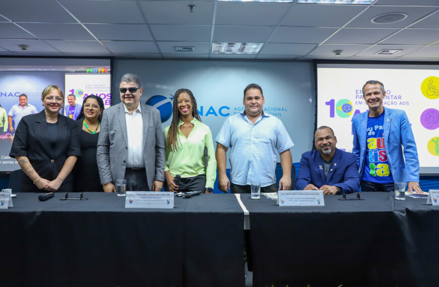 Sete pessoas diversas posam sorrindo em evento da ANAC, com telões e mesa à frente.