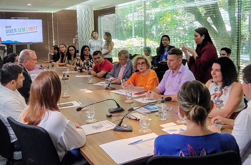 Reunião formal com várias pessoas em volta de mesa, tela ao fundo e intérprete de Libras.
