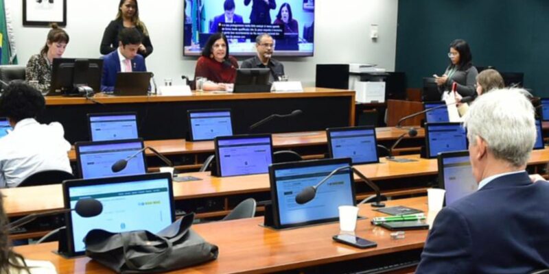 Plenário com pessoas em mesas, computadores, microfones e tela exibindo intérprete de Libras.