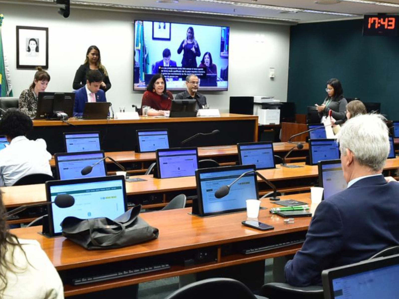 Plenário com pessoas em mesas, computadores, microfones e tela exibindo intérprete de Libras.
