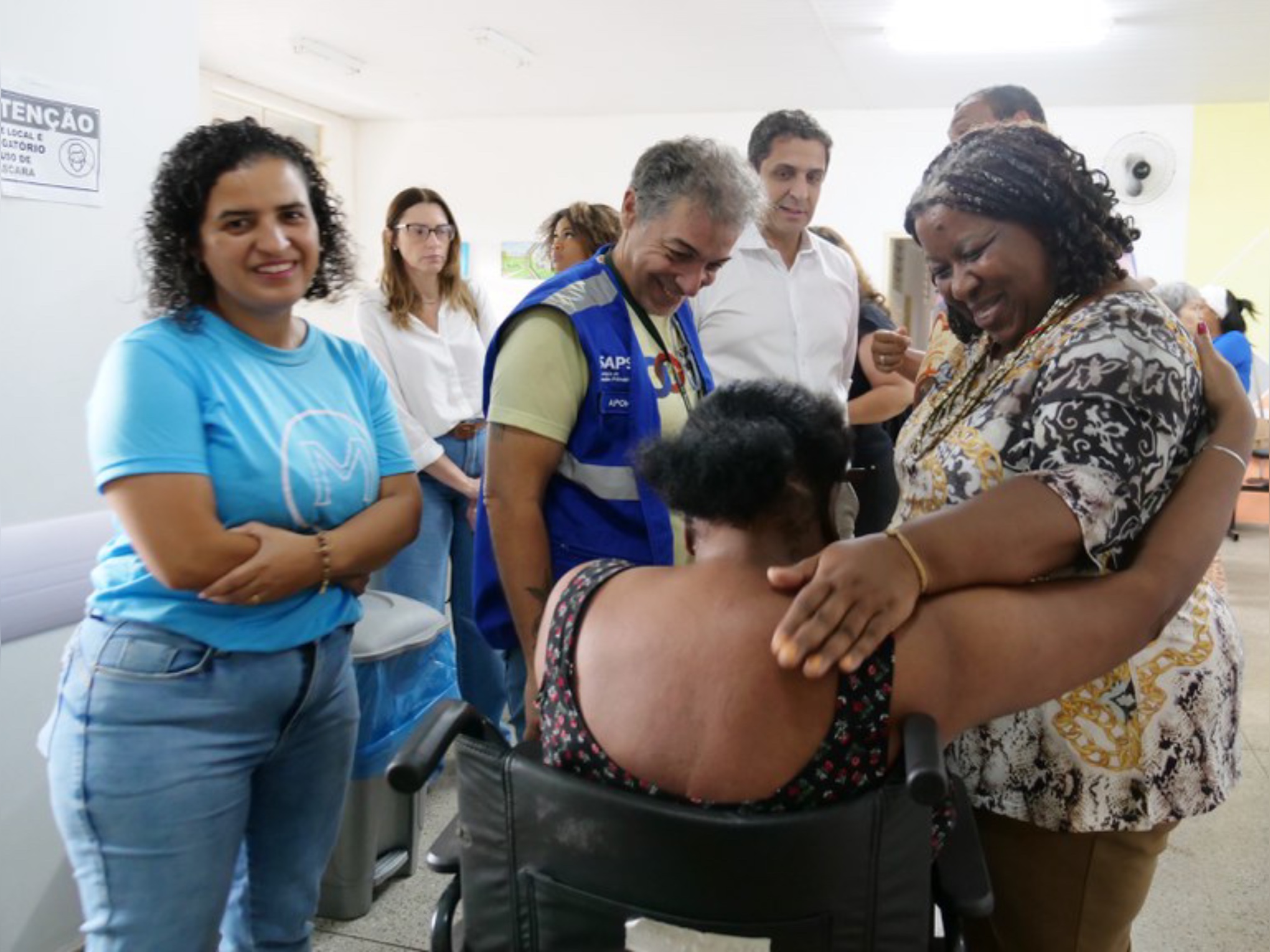 Grupo sorridente em clínica, mulher em cadeira de rodas recebe abraço acolhedor de outra mulher.