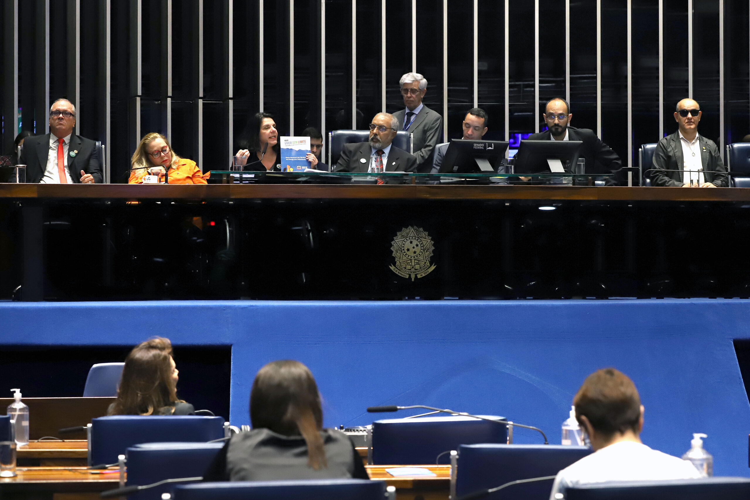 Sessão no Senado com autoridades na mesa diretora e pessoas assistindo ao fundo.