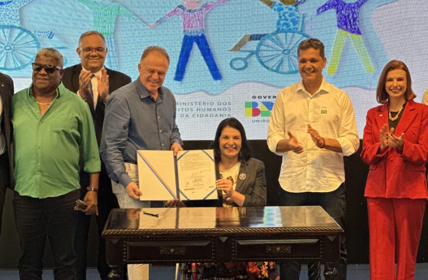 Grupo sorrindo em evento oficial, mulher em cadeira de rodas segura documento com homem.