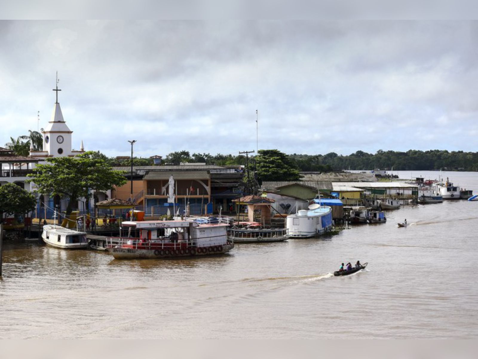 Margem de rio com barcos, casas simples, igreja com torre e pessoas em canoa na água barrenta.