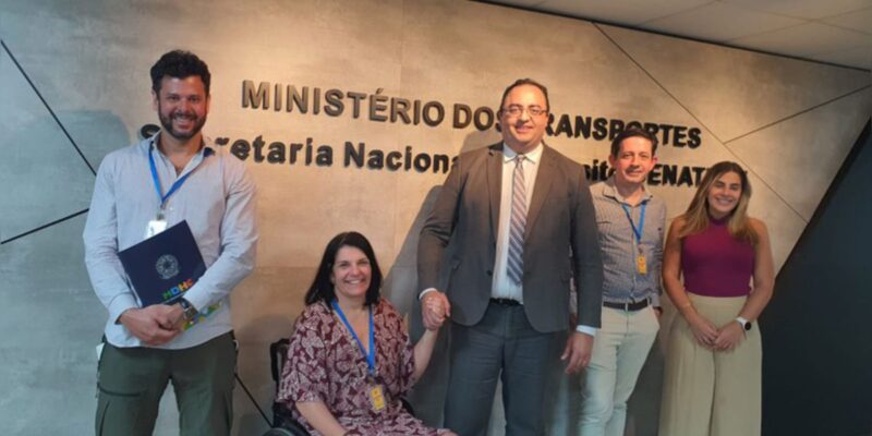 Cinco pessoas sorrindo em frente ao letreiro do Ministério dos Transportes, em ambiente formal.