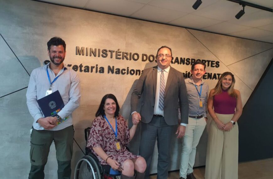 Cinco pessoas sorrindo em frente ao letreiro do Ministério dos Transportes, em ambiente formal.
