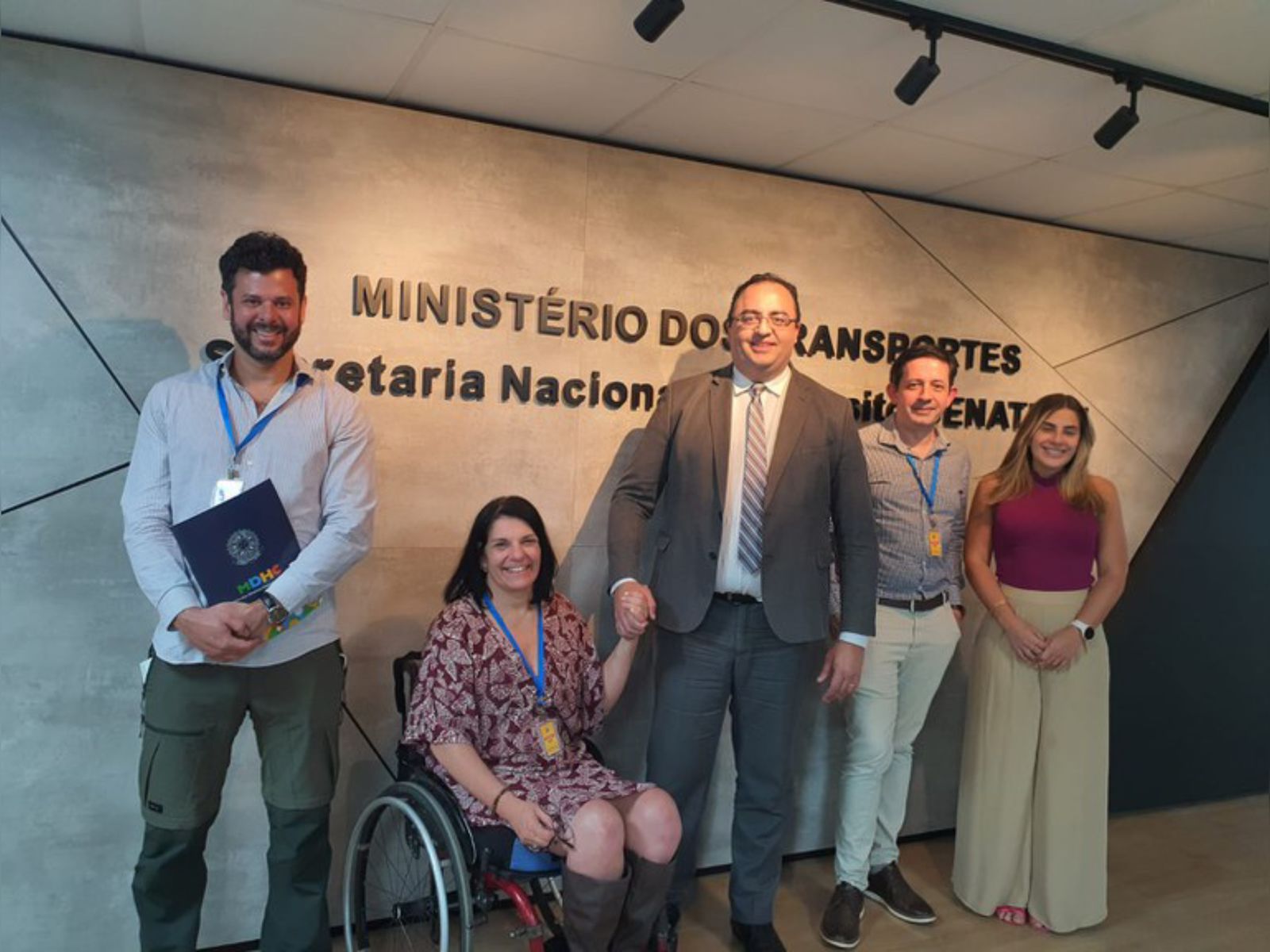 Cinco pessoas sorrindo em frente ao letreiro do Ministério dos Transportes, em ambiente formal.