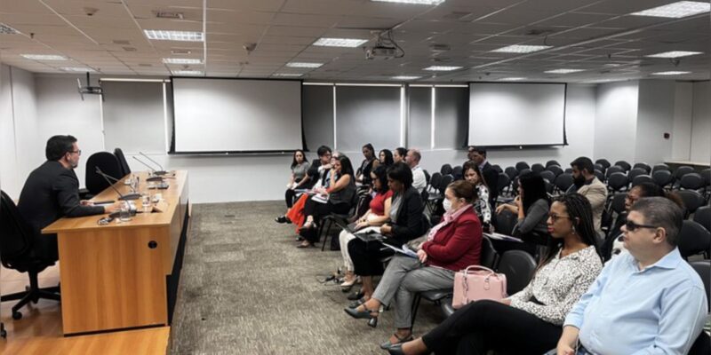 Auditório com pessoas atentas assistindo a um palestrante sentado à mesa, clima formal.