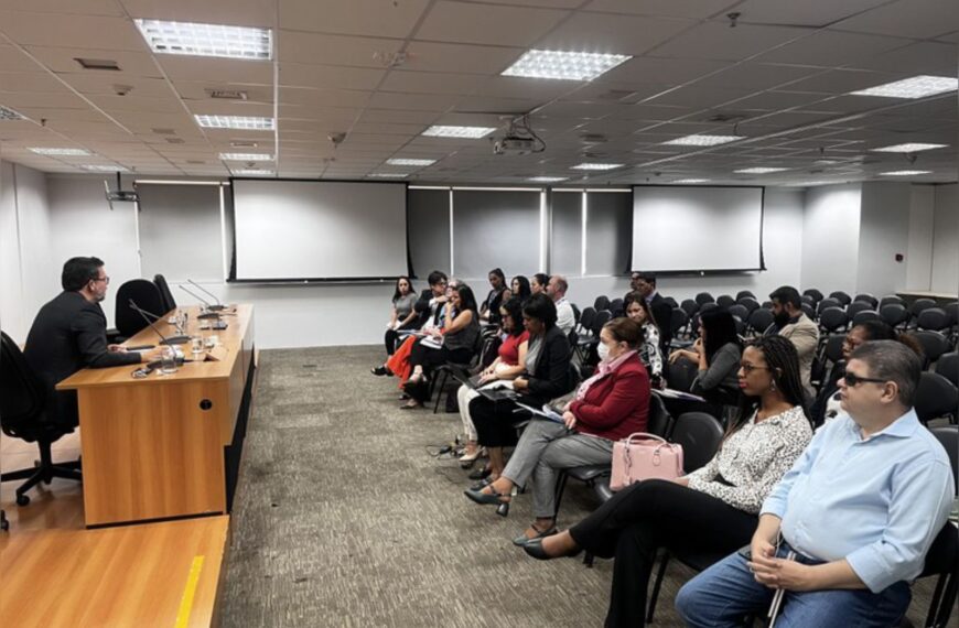 Auditório com pessoas atentas assistindo a um palestrante sentado à mesa, clima formal.