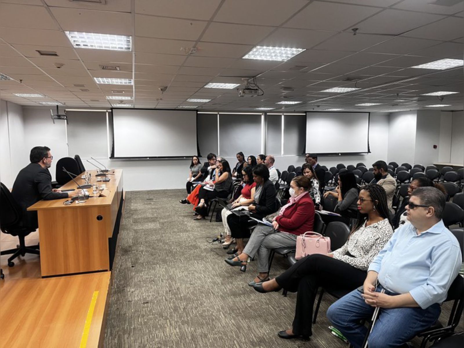 Auditório com pessoas atentas assistindo a um palestrante sentado à mesa, clima formal.