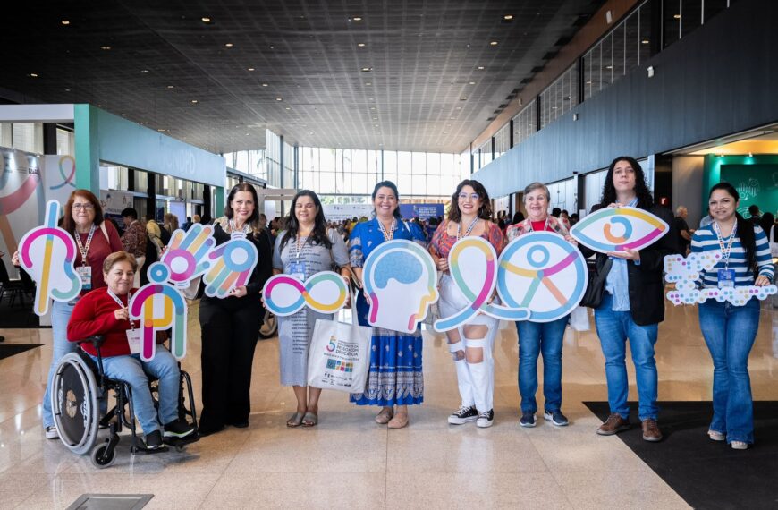Nove pessoas diversas, incluindo uma mulher cadeirante, seguram placas de inclusão em evento.