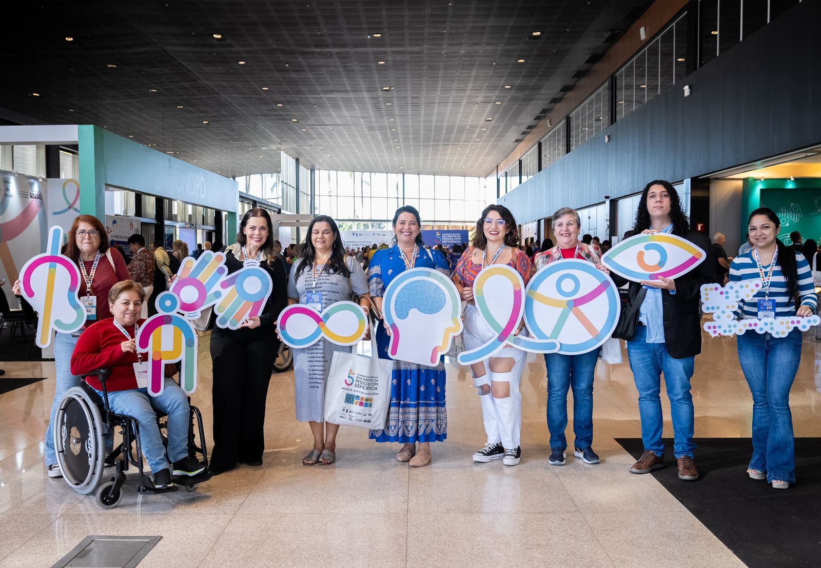 Nove pessoas diversas, incluindo uma mulher cadeirante, seguram placas de inclusão em evento.