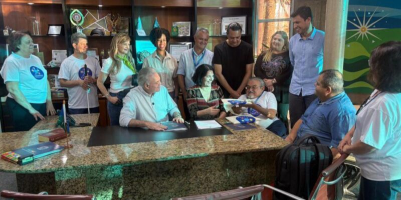Treze pessoas reunidas em escritório, sorrindo e interagindo ao redor de uma mesa.