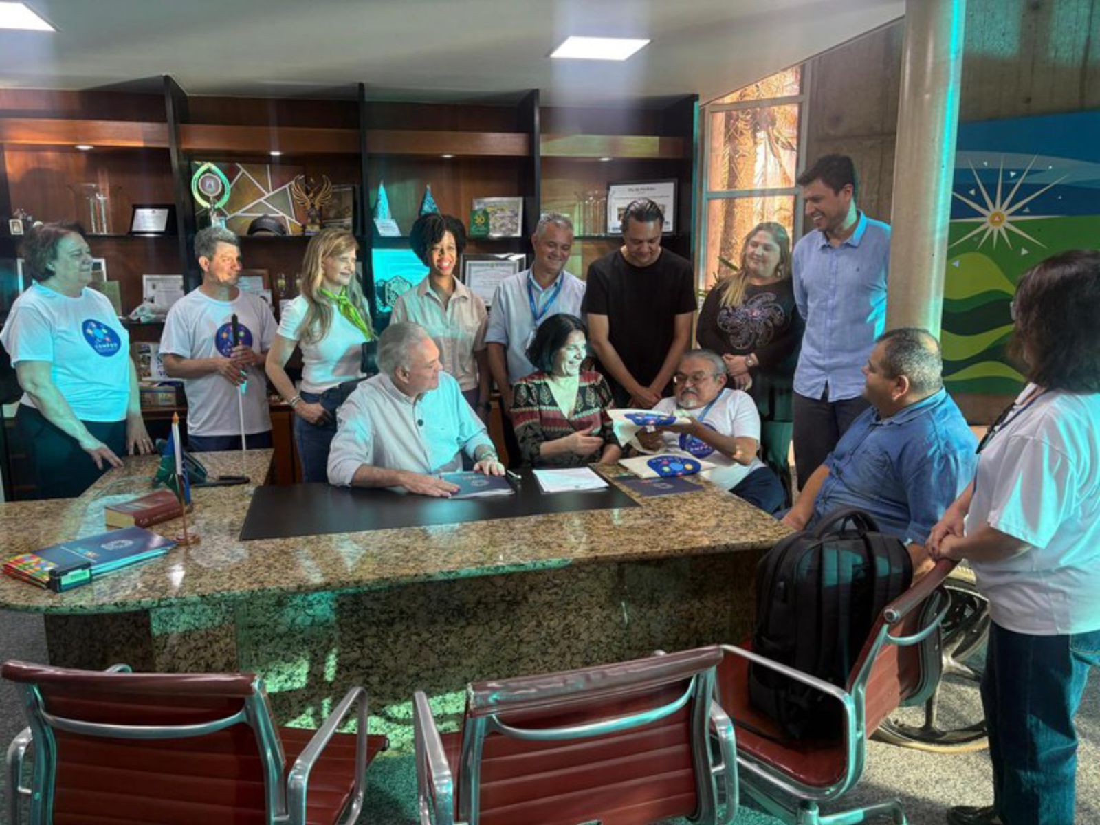 Treze pessoas reunidas em escritório, sorrindo e interagindo ao redor de uma mesa.