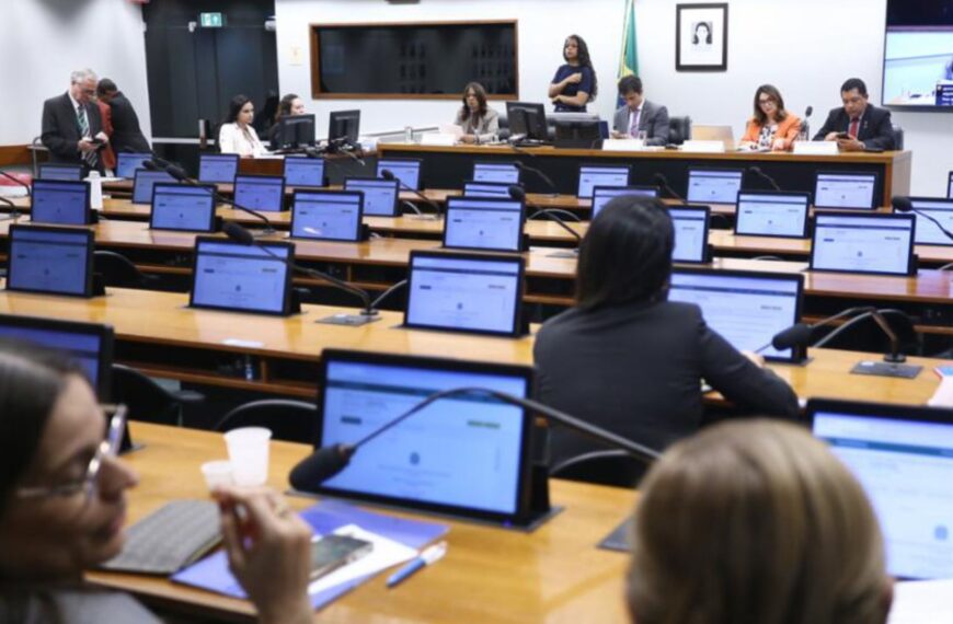 Sala de reunião parlamentar com mesas, monitores, microfones e pessoas em ambiente formal.