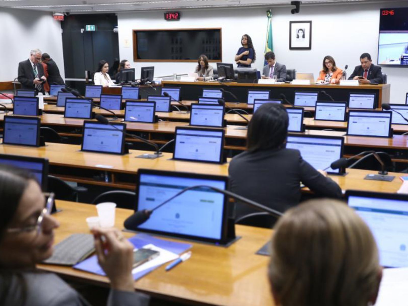 Sala de reunião parlamentar com mesas, monitores, microfones e pessoas em ambiente formal.