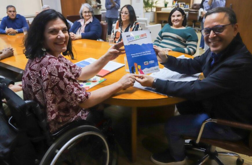 Grupo reunido, mulher em cadeira de rodas e homem mostram cartaz “Viver Sem Limite”.
