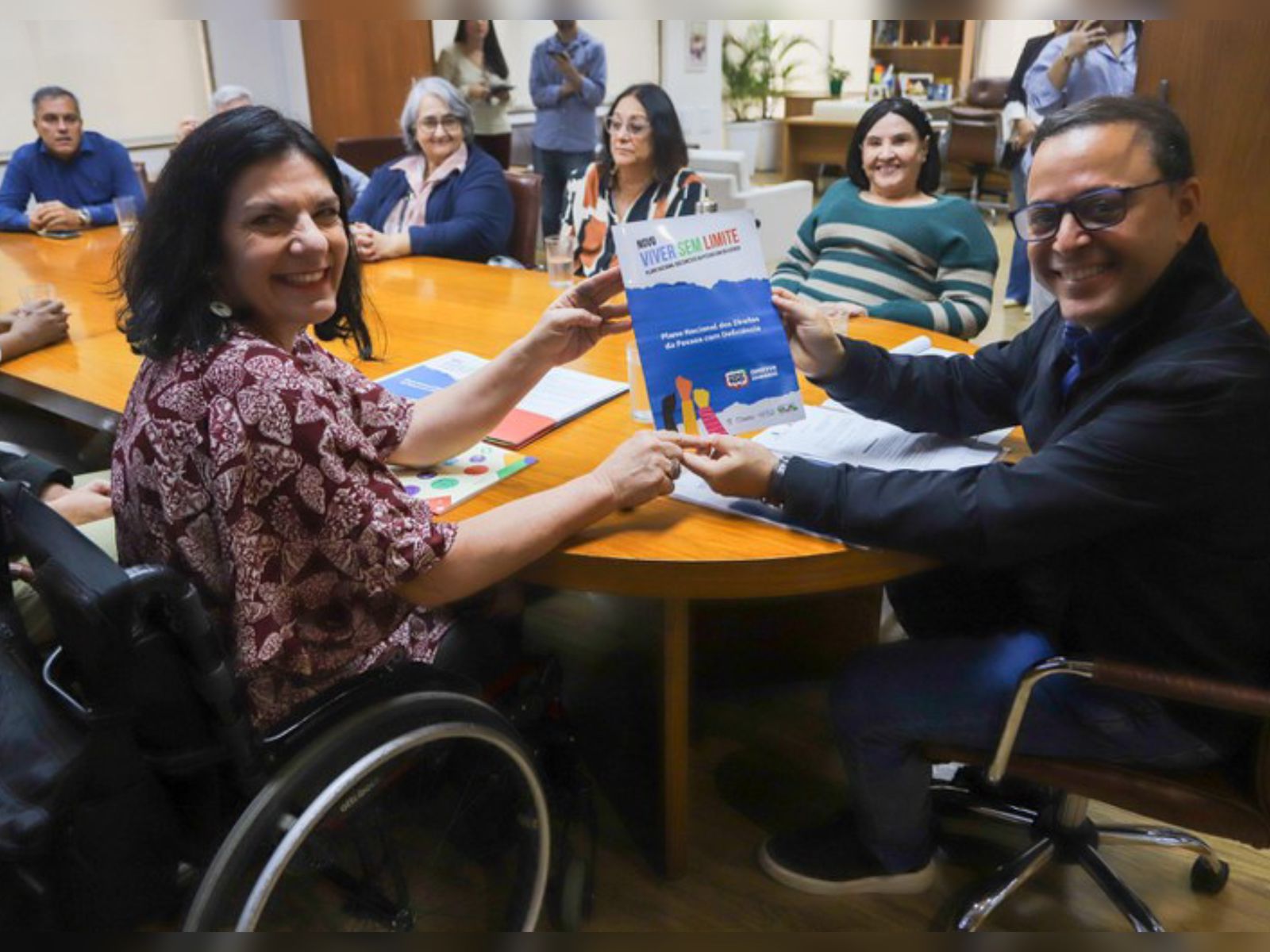 Grupo reunido, mulher em cadeira de rodas e homem mostram cartaz “Viver Sem Limite”.