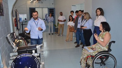 Homem apresenta equipamento; duas mulheres usuárias de cadeira de rodas acompanham a explicação.