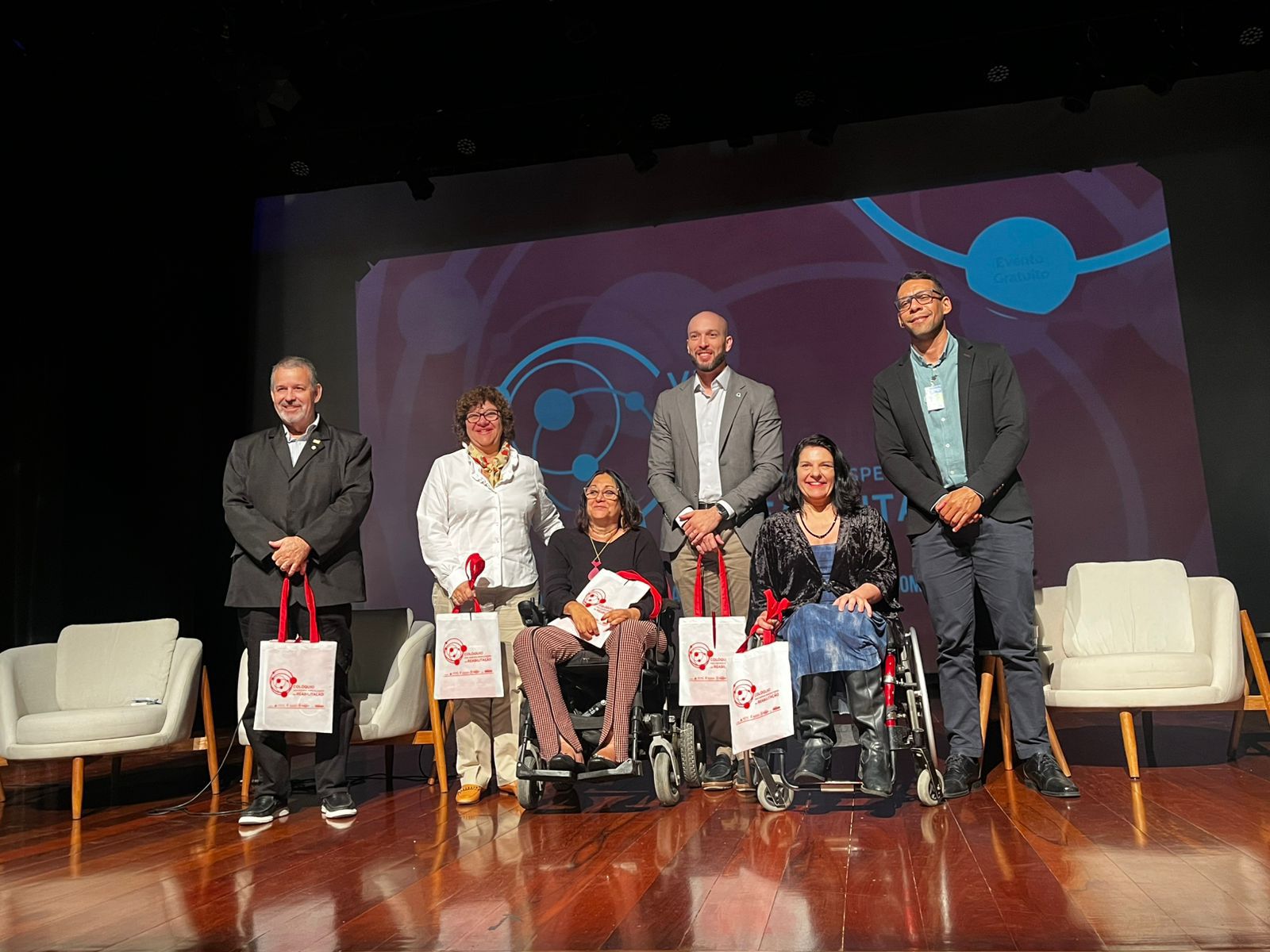 Seis pessoas sorrindo no palco, duas em cadeiras de rodas, segurando sacolas de brinde.