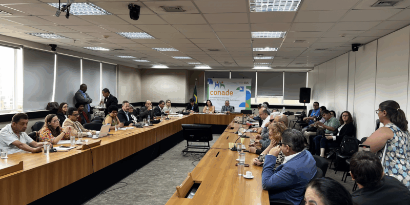 Sala ampla com mesas em U, pessoas reunidas em discussão, clima formal e profissional.