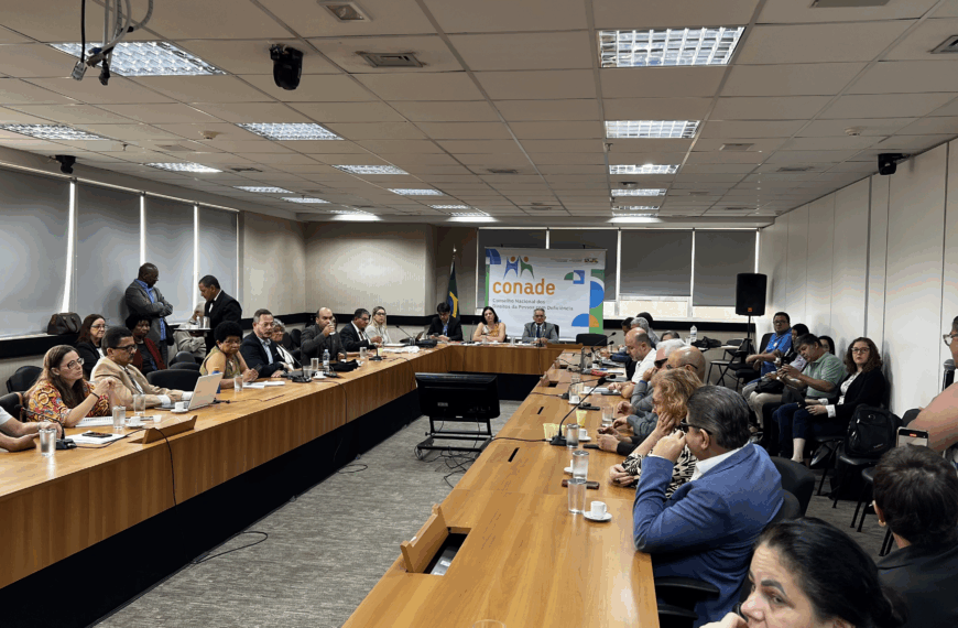 Sala ampla com mesas em U, pessoas reunidas em discussão, clima formal e profissional.