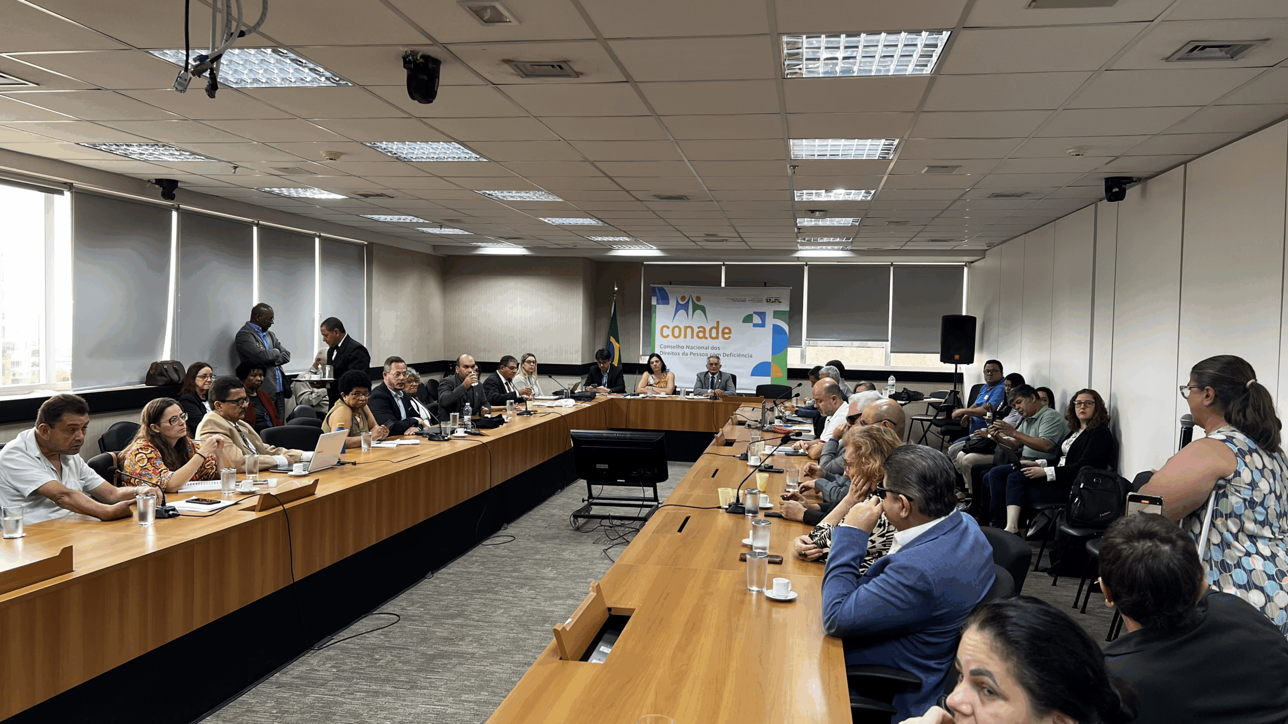 Sala ampla com mesas em U, pessoas reunidas em discussão, clima formal e profissional.