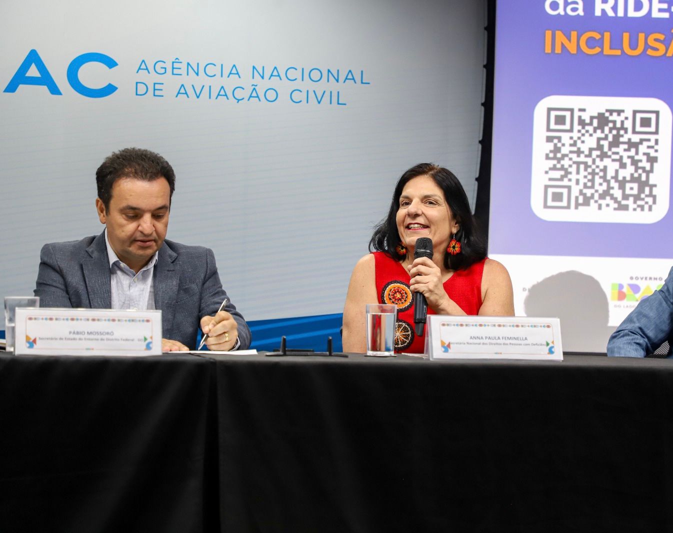 Homem e mulher em mesa de evento da ANAC; ela fala ao microfone, ambos com placas.