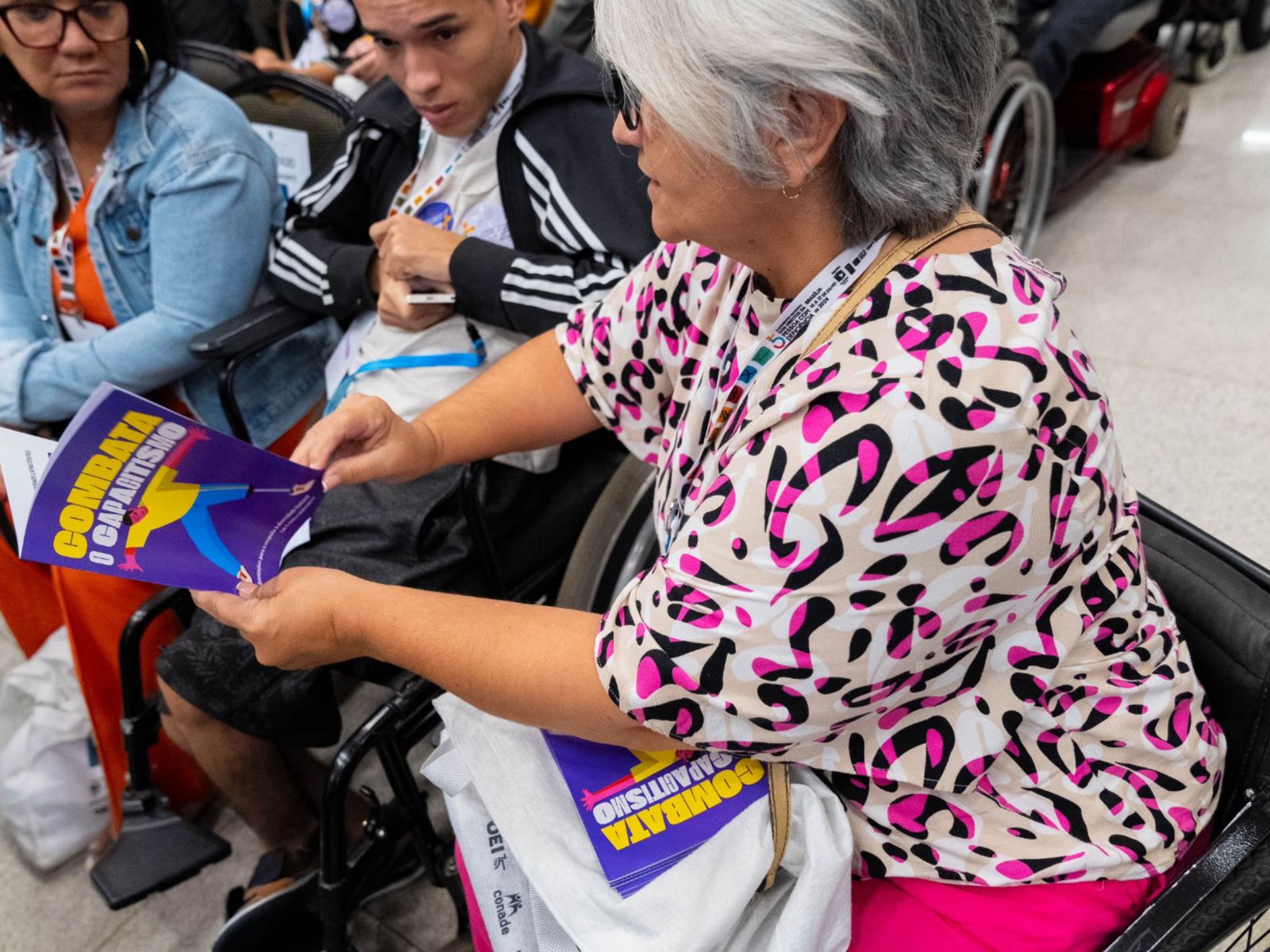 Mulher em cadeira de rodas lê livreto “Combata o Capitalismo” em evento com outras pessoas.