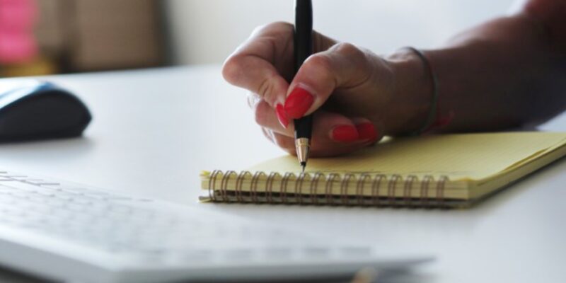 Mão feminina escreve em bloco amarelo com caneta preta, teclado e mouse ao fundo na mesa branca.