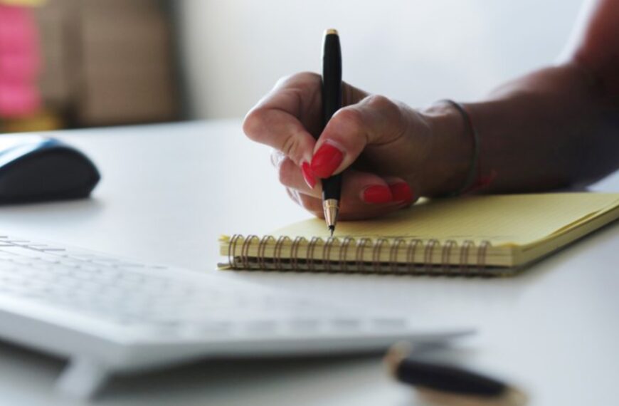 Mão feminina escreve em bloco amarelo com caneta preta, teclado e mouse ao fundo na mesa branca.