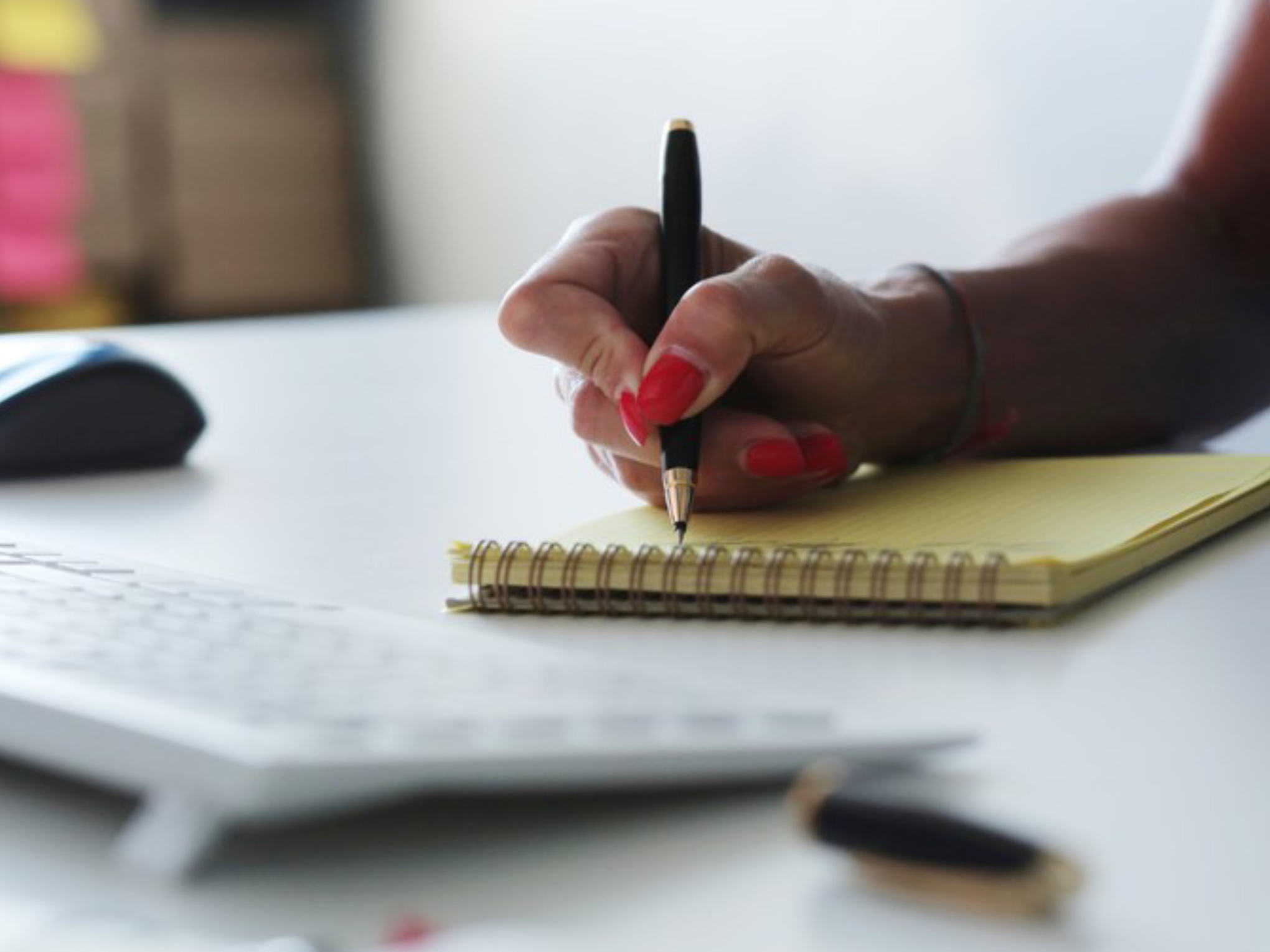 Mão feminina escreve em bloco amarelo com caneta preta, teclado e mouse ao fundo na mesa branca.