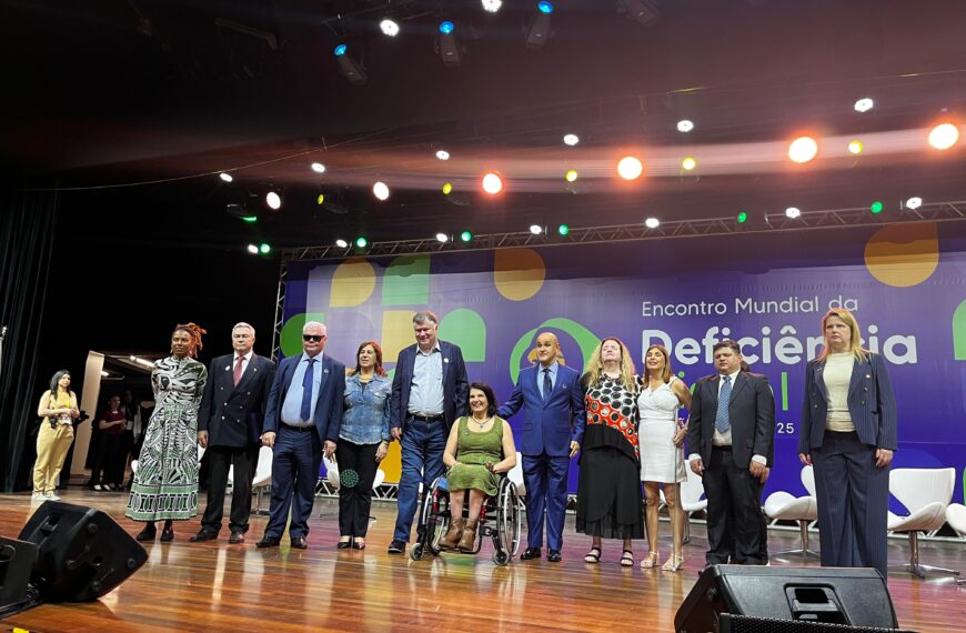 Doze pessoas diversas no palco do Encontro Mundial da Deficiência, com painel e luzes ao fundo.