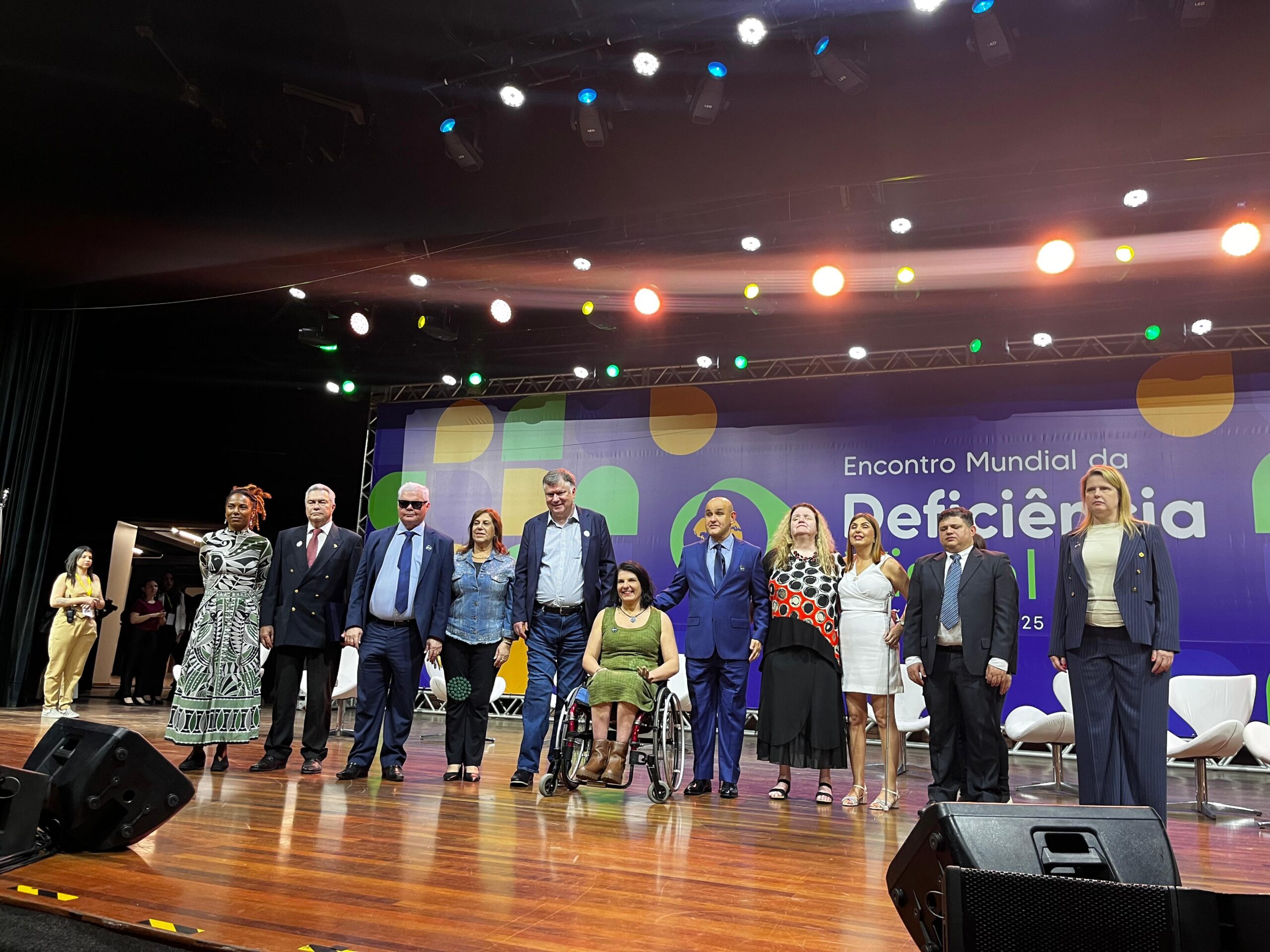 Doze pessoas diversas no palco do Encontro Mundial da Deficiência, com painel e luzes ao fundo.