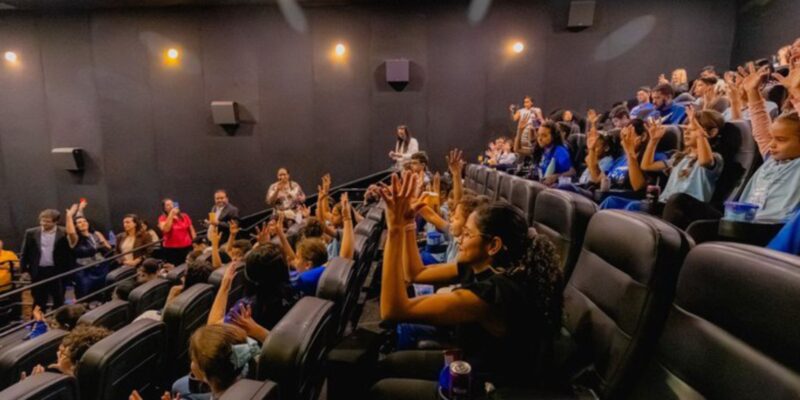 Crianças em sala de cinema com mãos levantadas e adultos observando, em ambiente animado.