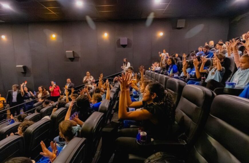 Crianças em sala de cinema com mãos levantadas e adultos observando, em ambiente animado.