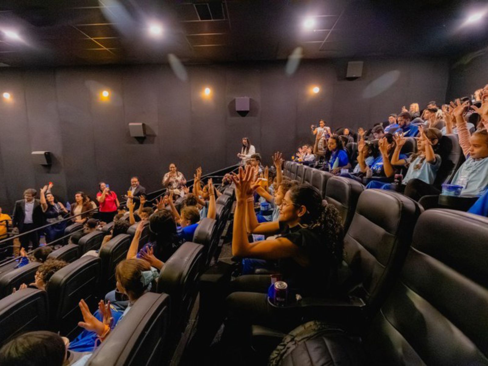 Crianças em sala de cinema com mãos levantadas e adultos observando, em ambiente animado.