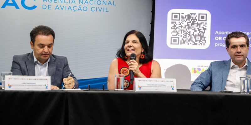 Três pessoas em mesa de evento, logo da ANAC e QR Code com “INCLUSÃO” ao fundo.
