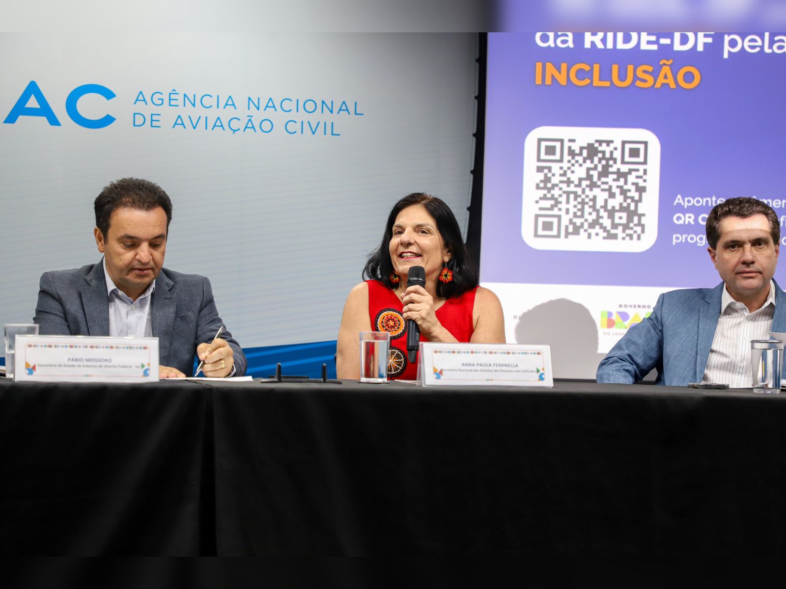 Três pessoas em mesa de evento, logo da ANAC e QR Code com “INCLUSÃO” ao fundo.