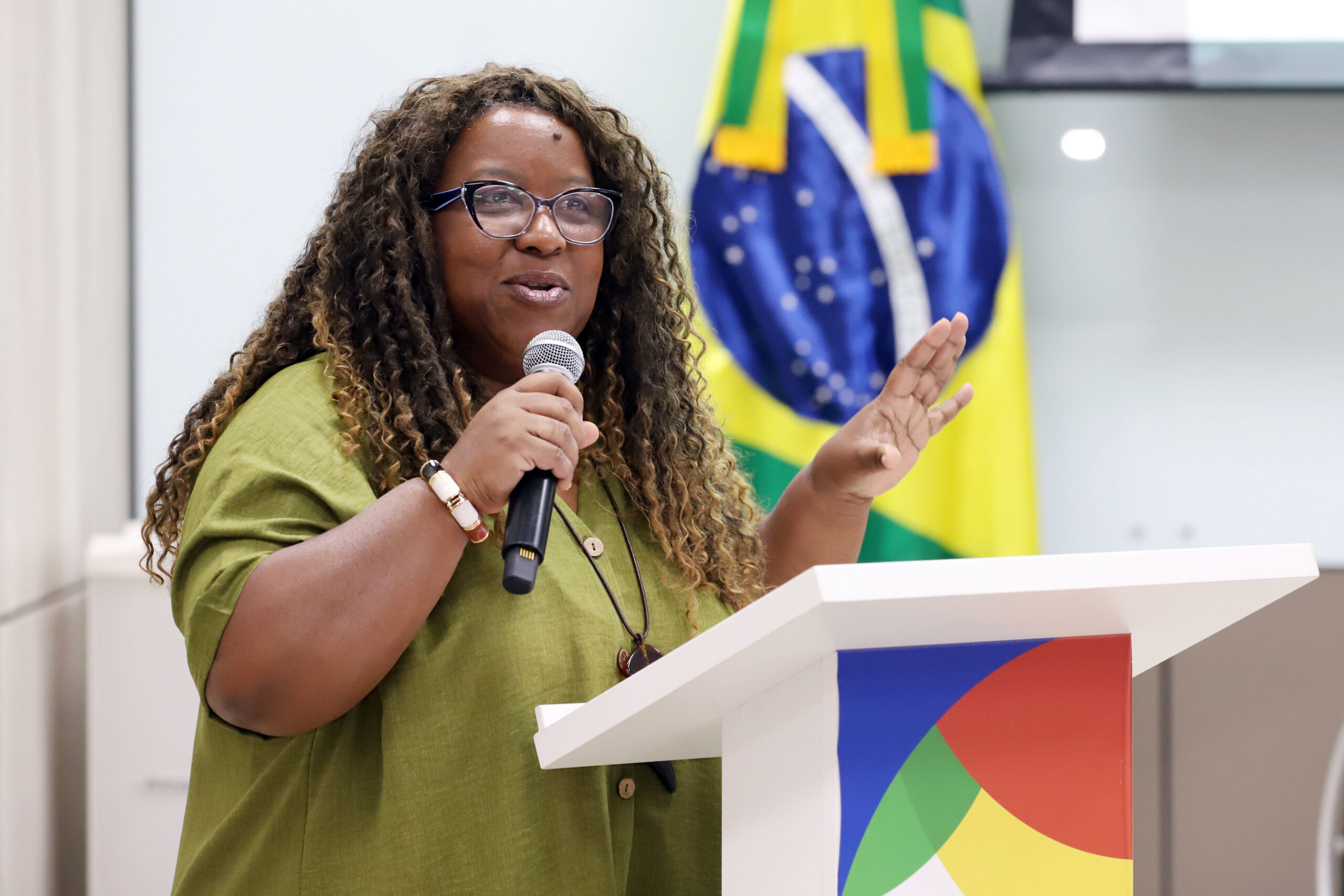 MDHC participa de lançamento da Rede Nacional de Pesquisa em Autismo e defende ciência para inclusão Ministra Macaé segura microfone em um púlpito, falando, com bandeira do Brasil ao fundo.